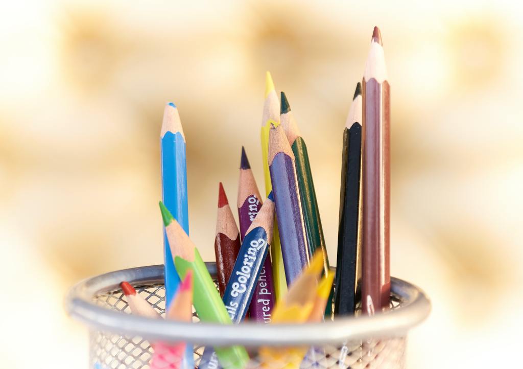 A desk tidy, half-filled with crayons of several colours and differing lengths that are standing upright or leaning at various angles. 